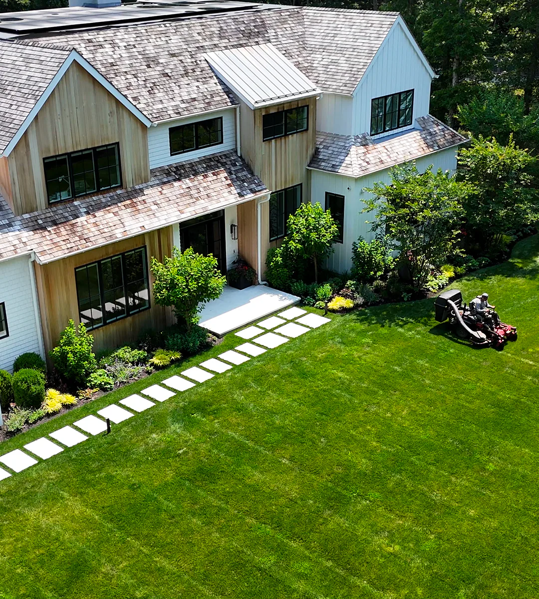 Aerial view of a Fenelon Landscapes crew member on a ride-on mower beside a shingled house with a stepping-stone path