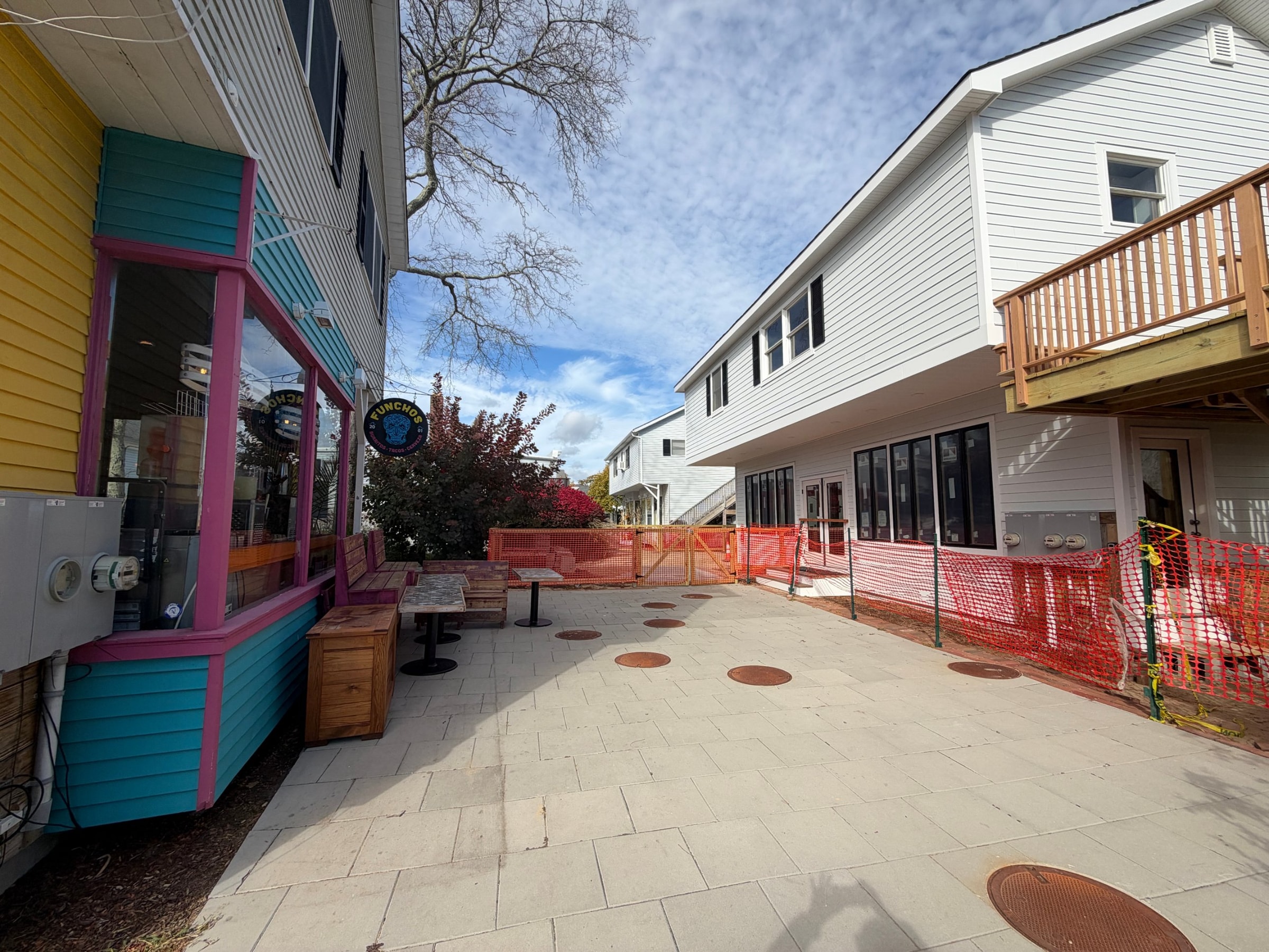 A paved courtyard beside the Funcho's building, with outdoor seating on the left and the new white building behind orange construction fencing on the right.