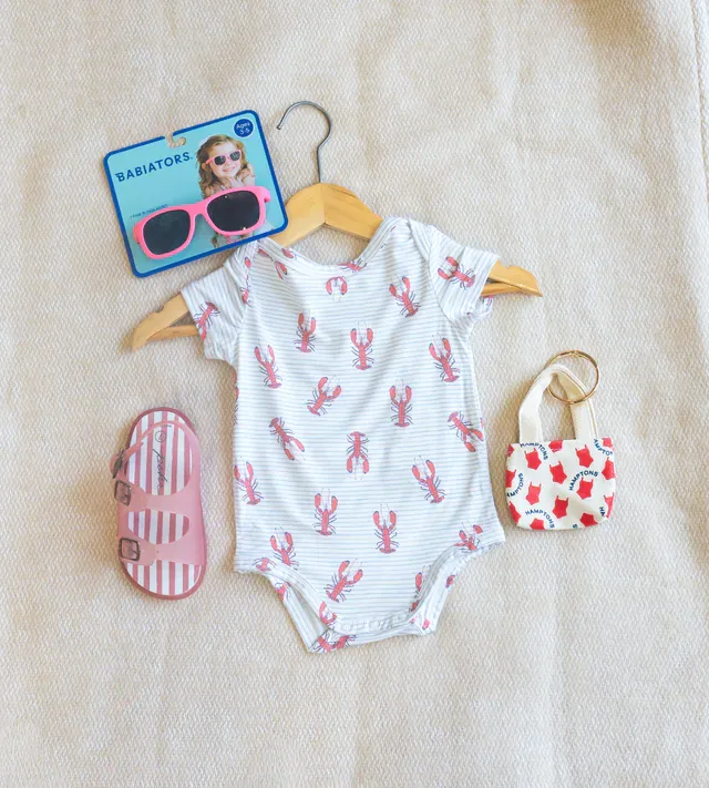 A lobster-print baby onesie on a wooden hanger with pink Babiators sunglasses, striped jelly sandals and a Hamptons mini tote, from Hildreth’s in Southampton