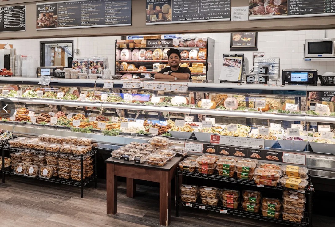 A deli counter with a Boar's Head menu board, a long case of salads and a team member behind the counter.