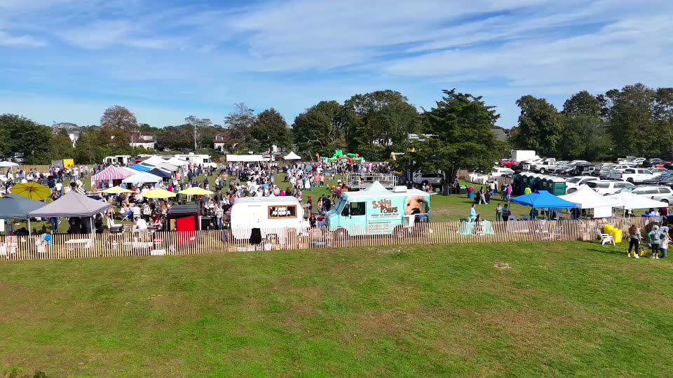 Aerial view of the Inlet to Inlet festival on the green in Westhampton Beach: tents, food trucks and a crowd behind a picket fence.